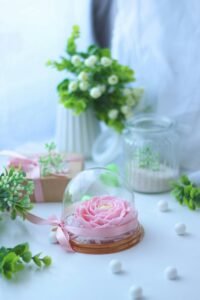 A beautiful pink flower arrangement under a glass cloche, surrounded by gifts and greenery in a serene setting.